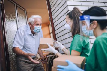 Young female volunteers in mask gives an elderly man boxes with food near his house. Quarantined, isolated. Coronavirus COVID-19. Donation