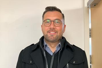 James Treadwell, a medium height man of approx. early 30s, with lightly tanned white skin and short hair styled in a small quiff, smiles warmly facing the camera. He is wearing smart casual outfit of an open-necked checkered shirt with v-neck jumper, both in blue, and a black casual parka. He is wearing brown framed glasses and a black lanyard carrying a brightly coloured ID badge. He is photographed against a cream background similar to a public building interior wall.    